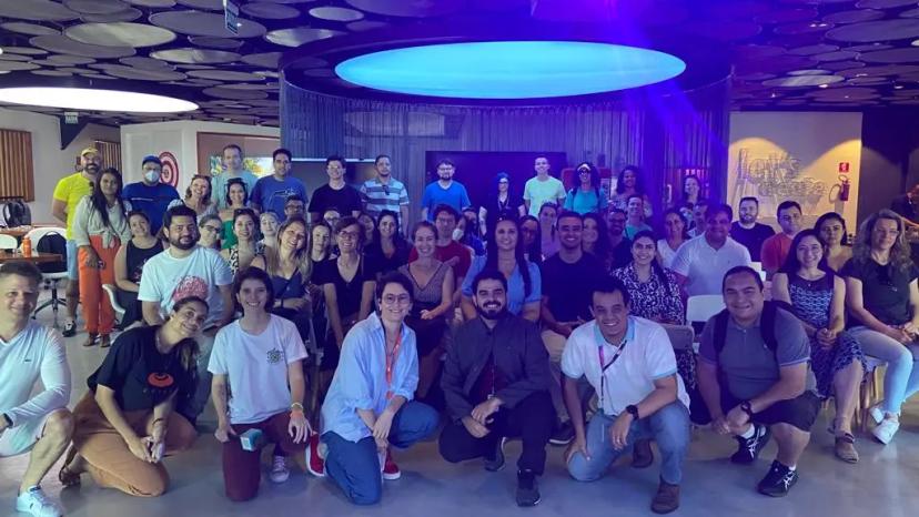Bootcamp Banco do Brasil – a transição estratégica do remoto ao presencial Uma grande equipe de profissionais posa reunida para uma foto coletiva em um ambiente interno moderno e amplo, iluminado por uma grande estrutura circular de luz azul no teto