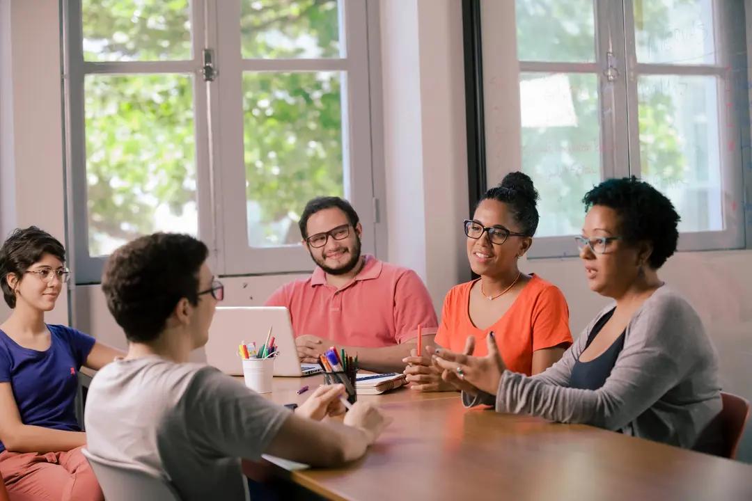 Design expansivo – inovação e soluções através da construção coletiva Grupo de cinco pessoas reunidas em torno de uma mesa de madeira em um escritório iluminado por janelas ao fundo, enquanto conversam e interagem de forma colaborativa