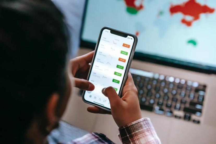 Banking as a Service – impactos, vantagens e inovação nos negócios Pessoa segura um celular que exibe gráficos e valores financeiros, com a tela de um notebook ao fundo