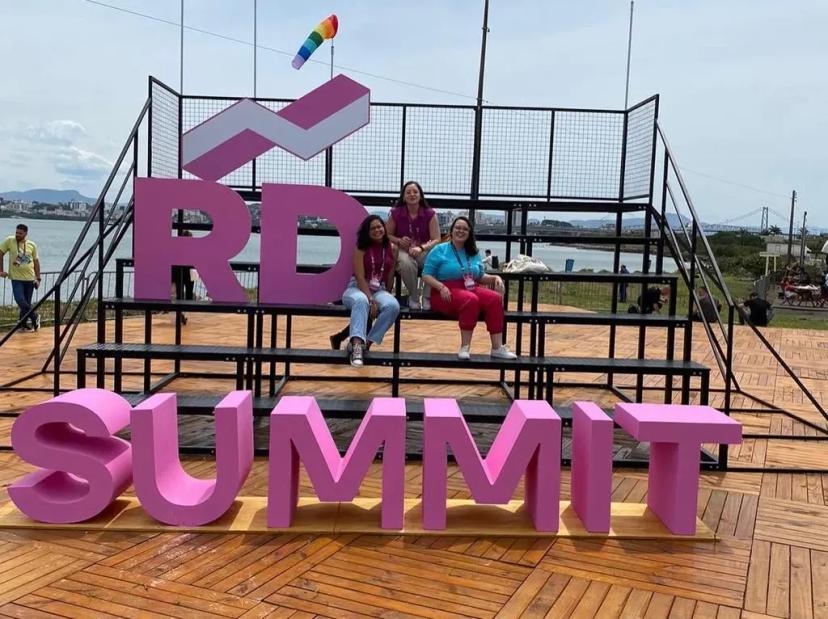 Inovação em marketing – lições e insights no RD Summit 2022 Três mulheres sorriem sentadas em uma estrutura de arquibancada montada em uma área externa com piso de madeira, posicionadas logo atrás de um grande letreiro tridimensional rosa que forma a palavra "SUMMIT" e abaixo das letras "RD"
