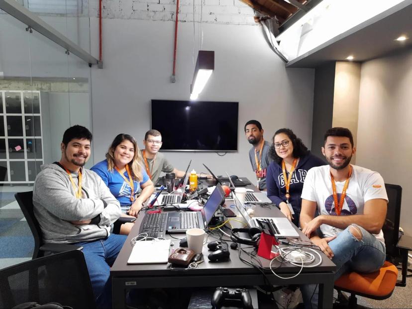 Code Bootcamp – como a capacitação prática otimizou a automação de testes no CESAR Grupo de seis pessoas sorri sentando ao redor de uma grande mesa de escritório cheia de notebooks, cabos e canecas, voltadas para a foto em uma sala de reuniões