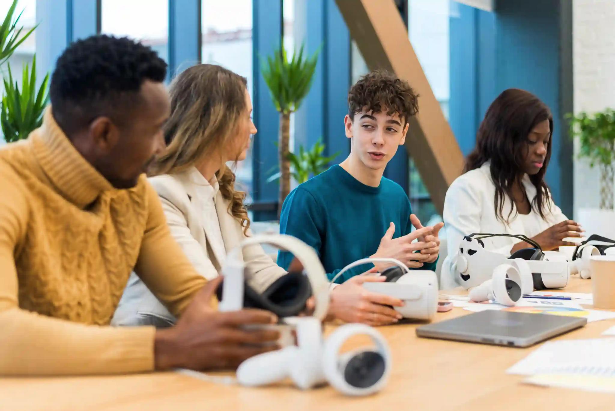 Hana Electronics e CESAR – Formação em IoT para a Amazônia Ocidental Quatro jovens em reunião numa mesa com óculos de realidade virtual. Um rapaz explica ideias enquanto os outros ouvem.