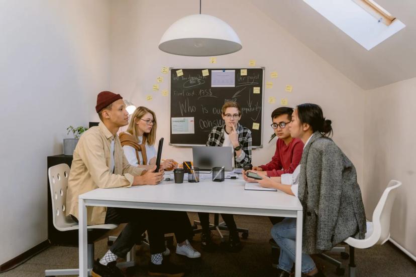 Primeira liderança técnica – 3 dicas essenciais para começar sua jornada Cinco jovens profissionais estão reunidos ao redor de uma mesa branca de escritório em uma sala de reuniões, conversando e debatendo projetos diante de um quadro-negro com anotações em giz ao fundo