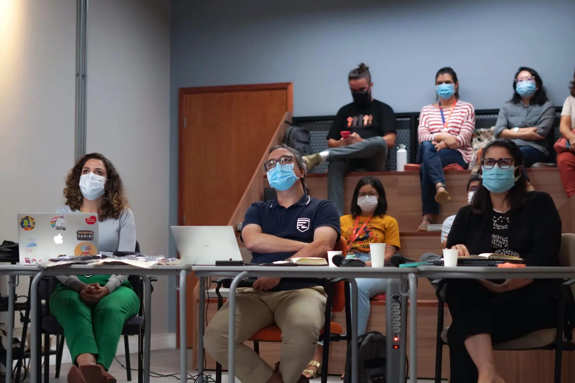 Intraempreendedorismo e Inovação Corporativa Três profissionais sentados à mesa com notebooks e outras pessoas ao fundo em uma arquibancada assistindo a um treinamento.