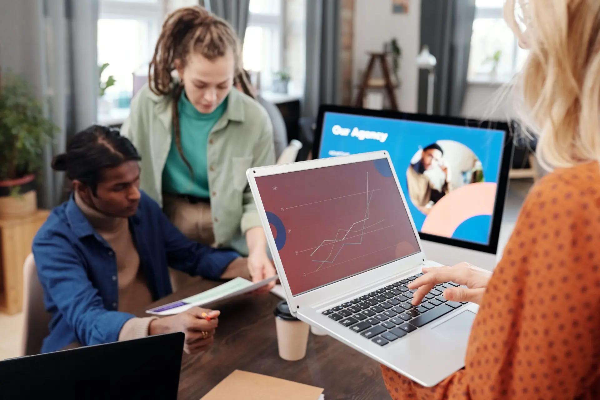 Alinhamento estratégico e colaboração através do Design Driven Três pessoas analisam gráficos em um laptop e monitor em escritório. Uma mulher digita enquanto colegas observam papéis.