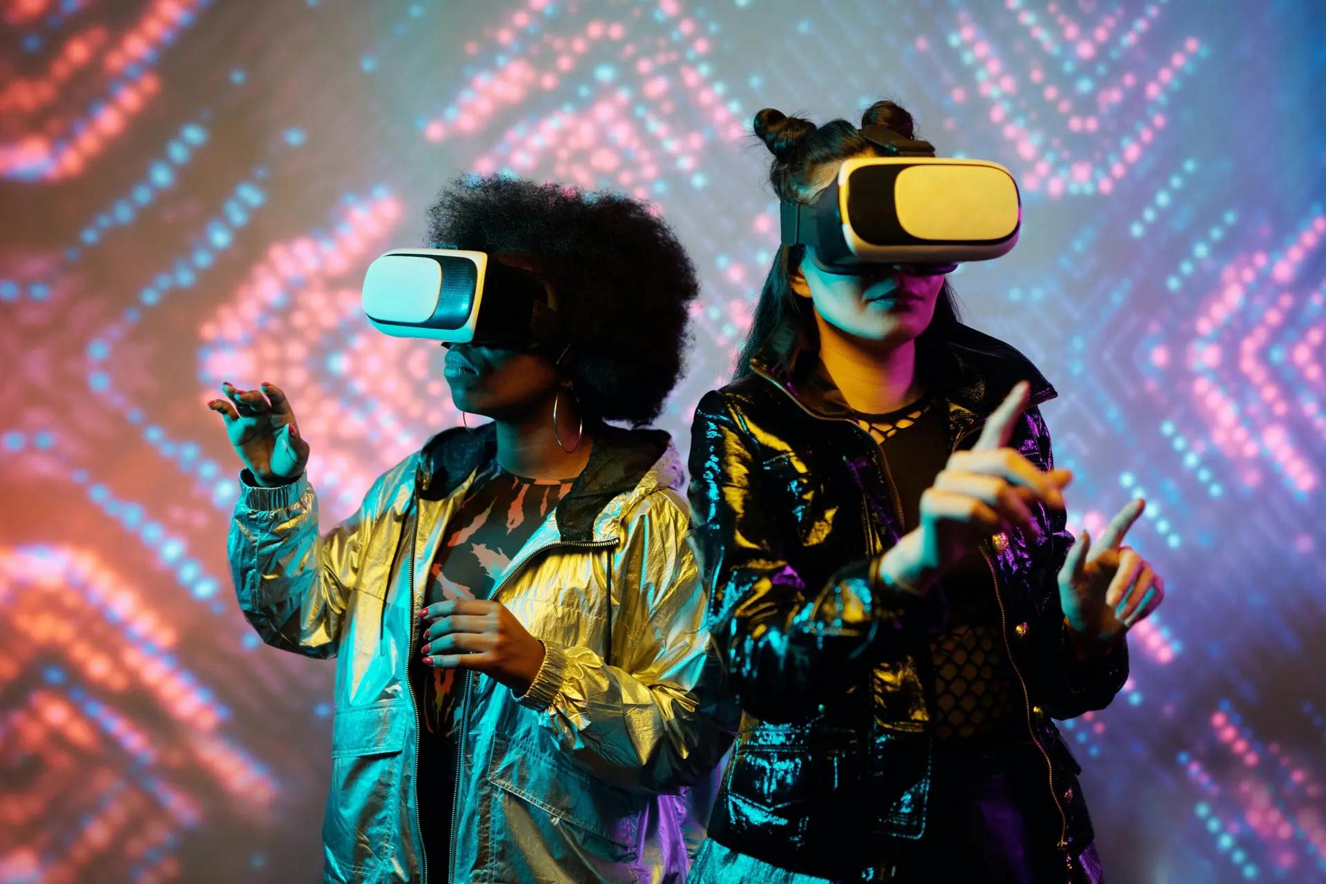 Designer do Metaverso – Criando Experiências Imersivas no Mundo Virtual Duas mulheres usando óculos de realidade virtual e roupas metalizadas, gesticulando em frente a um fundo de luzes neon.