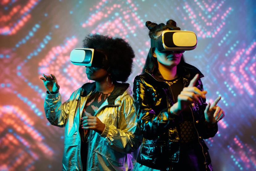 Designer do Metaverso – Criando Experiências Imersivas no Mundo Virtual Duas mulheres usando óculos de realidade virtual e roupas metalizadas, gesticulando em frente a um fundo de luzes neon.