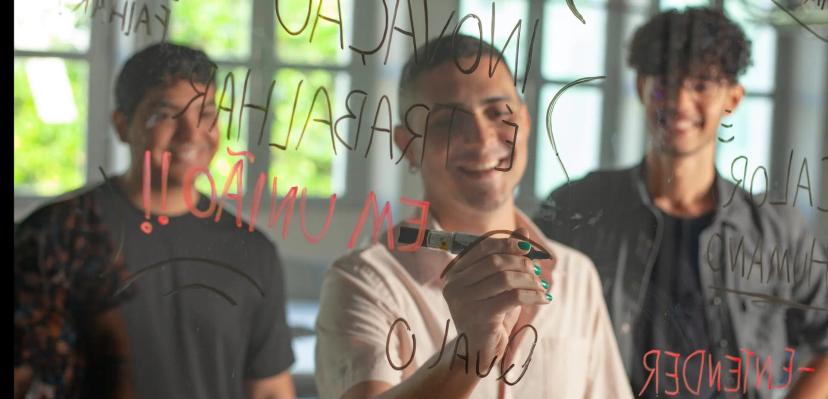 Intraempreendedor e as características do inovador corporativo Homem com caneta escreve em painel de vidro transparente palavras como inovação e união, ladeado por dois colegas sorridentes.