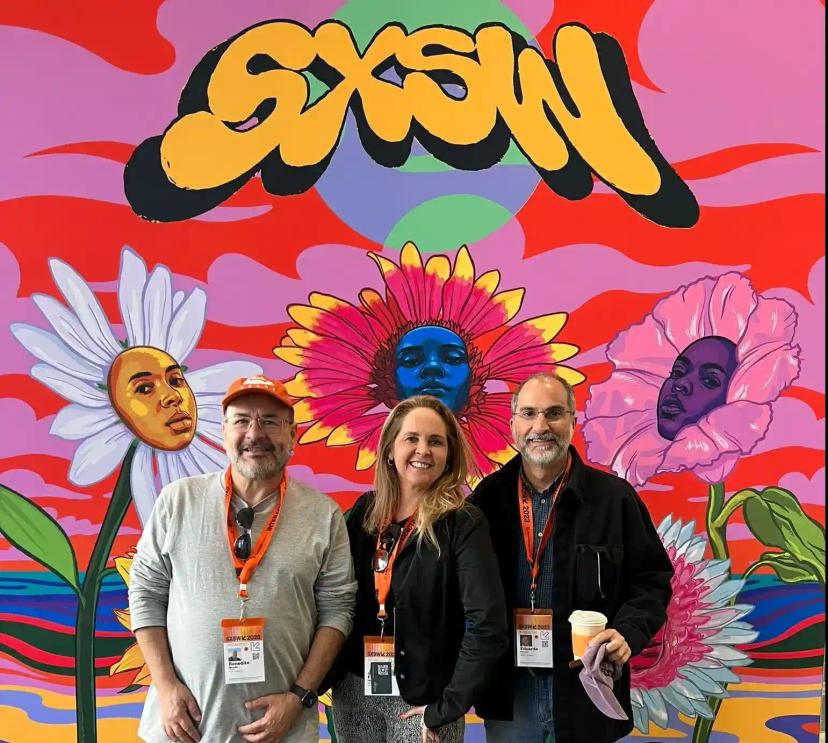 SXSW 2023 – Inovação e tendências globais com o time do CESAR. Três pessoas sorrindo em frente a um painel colorido com flores e a logo SXSW, usando crachás do evento em Austin.