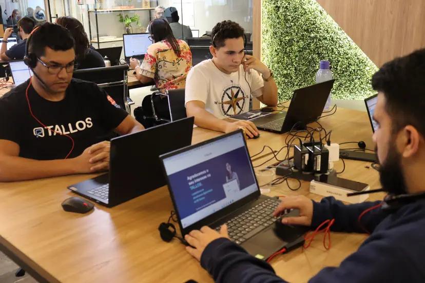Tallos utiliza tecnologia de ponta para impulsionar startups Três homens trabalham com notebooks em mesa de madeira. Um deles veste camisa preta com o logo da Tallos e usa headset.