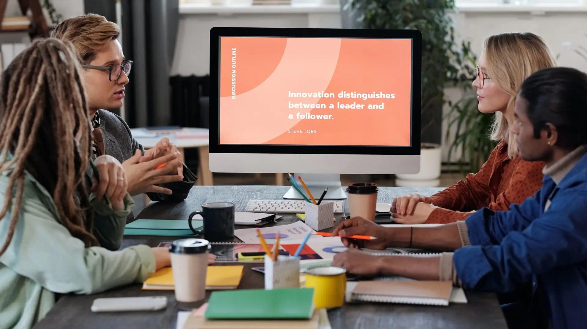 Gestão da inovação – o que é e como aplicar na sua empresa Grupo de quatro pessoas em reunião ao redor de mesa com café, observando monitor com a frase sobre liderança e inovação