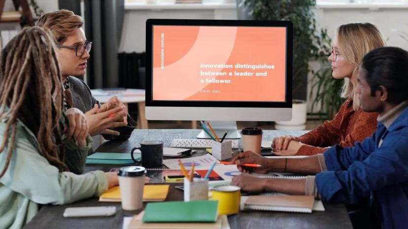 Gestão da inovação – o que é e como aplicar na sua empresa Grupo de quatro pessoas em reunião ao redor de mesa com café, observando monitor com a frase sobre liderança e inovação
