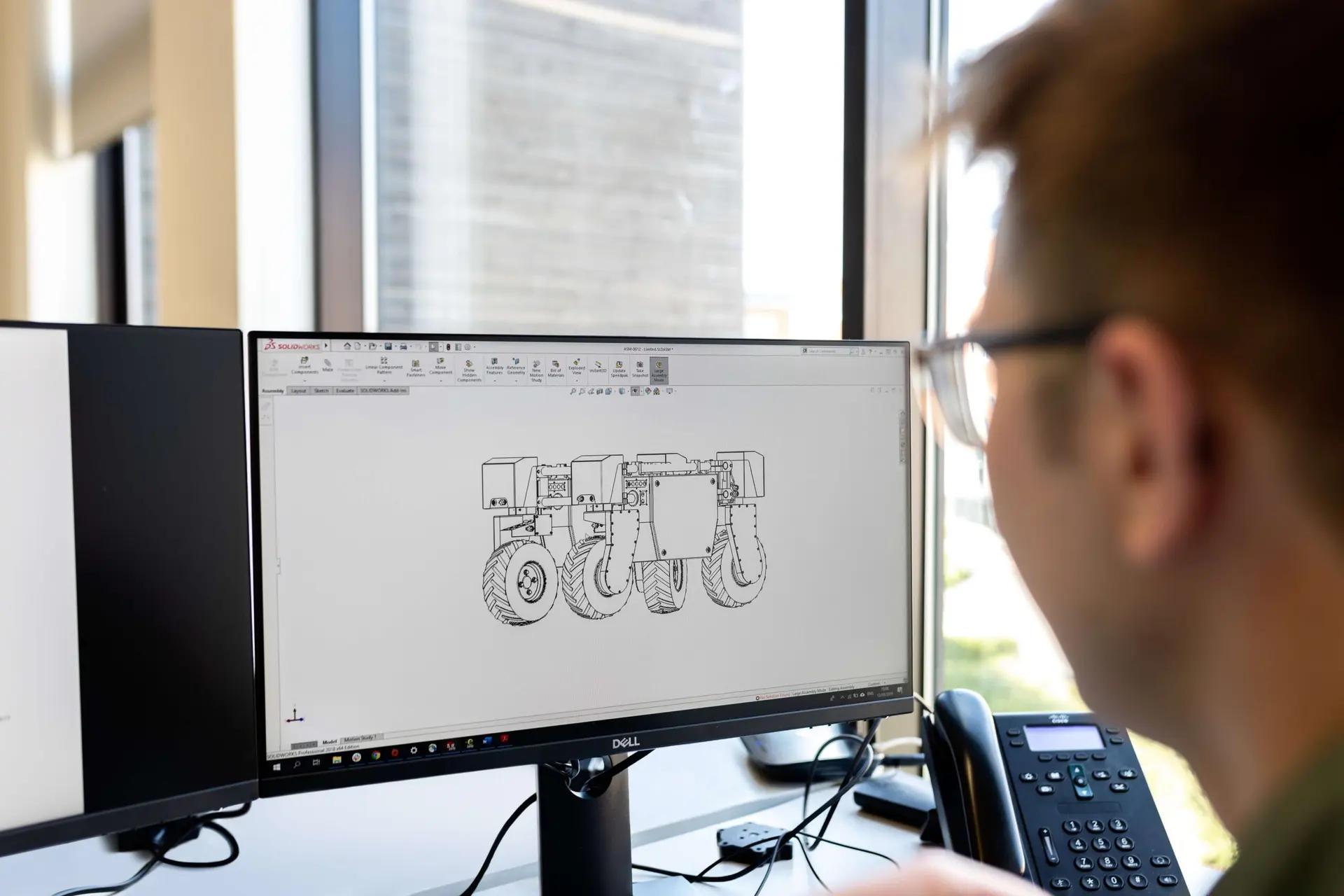 Indústria 4.0 – Tecnologias e o impacto na manufatura avançada Homem de óculos observa monitor exibindo software CAD com desenho técnico 3D de um veículo robótico de seis rodas.