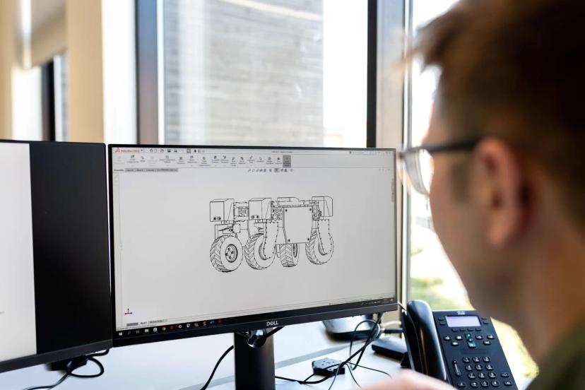 Indústria 4.0 – Tecnologias e o impacto na manufatura avançada Homem de óculos observa monitor exibindo software CAD com desenho técnico 3D de um veículo robótico de seis rodas.