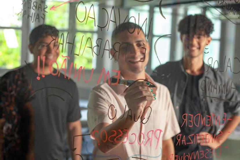 Design Thinking aplicado na resolução de problemas e inovação Homem escreve em painel de vidro transparente durante dinâmica de Design Thinking com dois colegas sorrindo ao fundo.