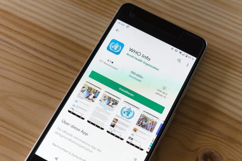 Healthtechs e o impacto das tecnologias na saúde e sociedade Smartphone sobre mesa de madeira exibe a loja de aplicativos com o app oficial da Organização Mundial da Saúde (OMS).