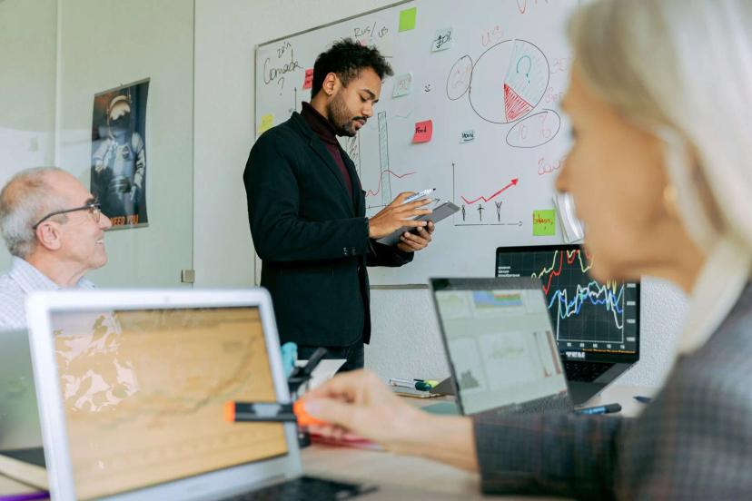 Inovação radical – estratégias para transformar o mercado atual Homem jovem desenha gráficos em quadro branco enquanto colegas analisam dados em laptops em um escritório de tecnologia.