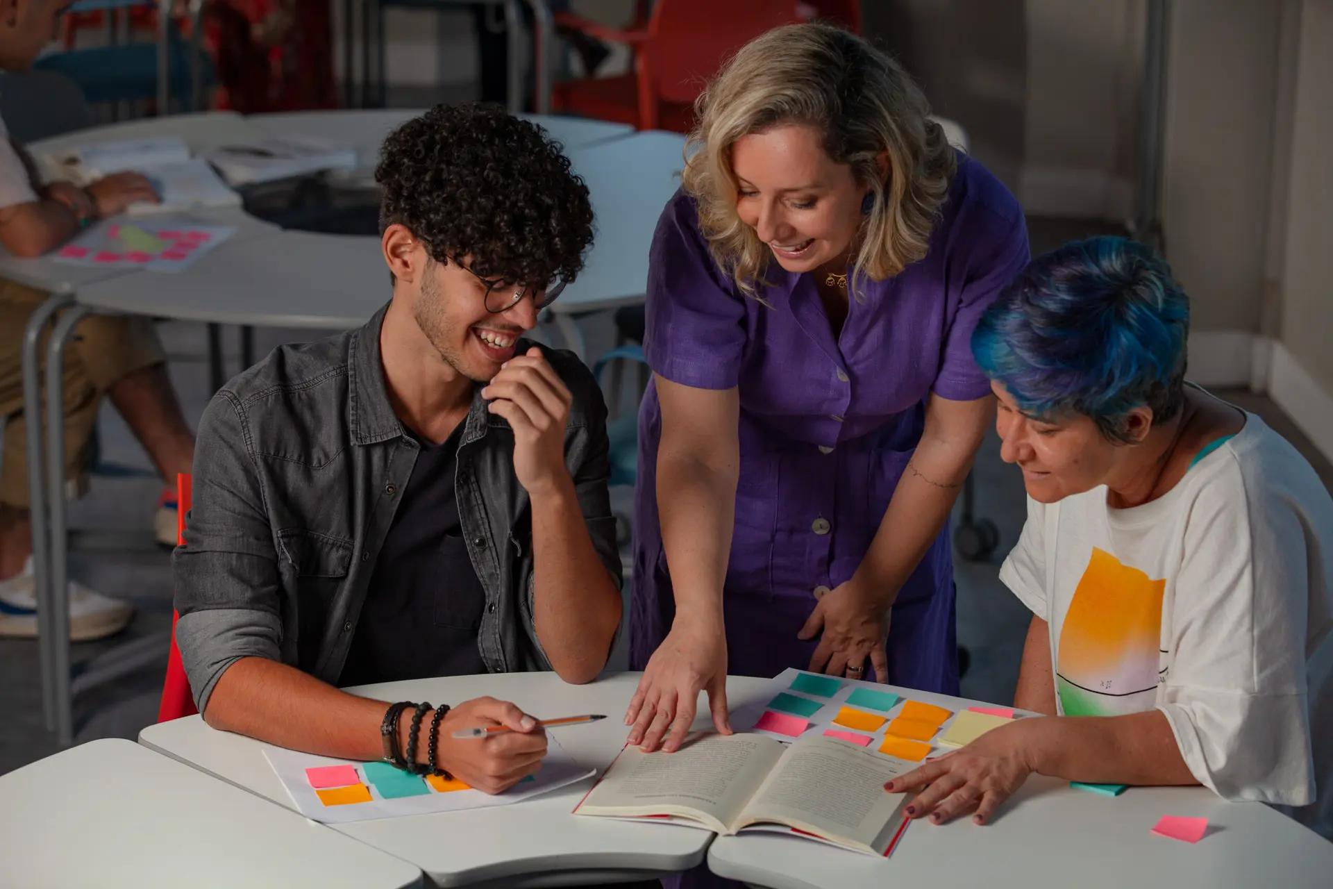 Lideranças em projetos de inovação e seu papel estratégico Três pessoas sorriem ao redor de uma mesa com um livro aberto e vários post-its coloridos em uma dinâmica de liderança.