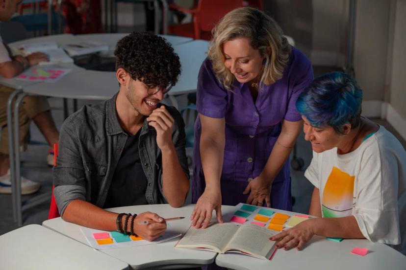 Lideranças em projetos de inovação e seu papel estratégico Três pessoas sorriem ao redor de uma mesa com um livro aberto e vários post-its coloridos em uma dinâmica de liderança.