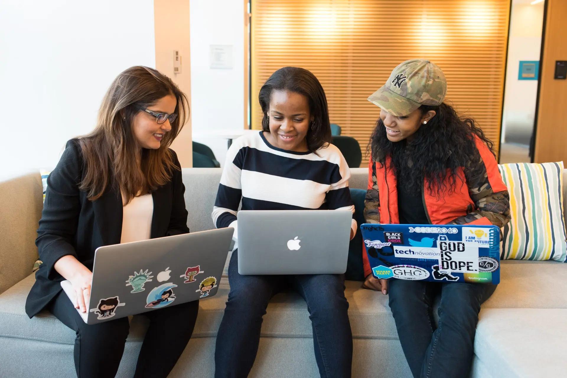 Lifelong learning nas empresas e o desenvolvimento contínuo de talentos Três mulheres sorridentes sentadas em um sofá, cada uma com um laptop, colaborando em um ambiente moderno de escritório.