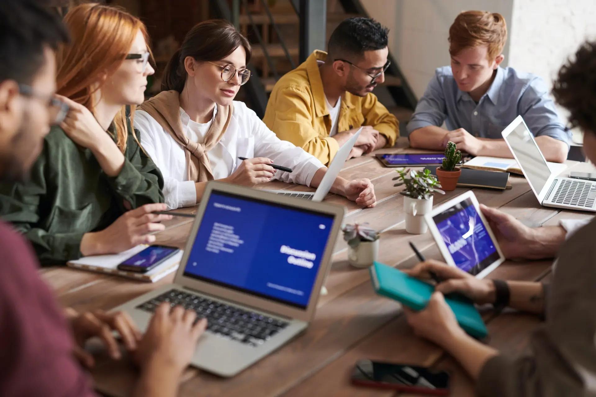 Soluções digitais inovadoras – os 4 pilares essenciais para o sucesso Grupo de sete pessoas em reunião ao redor de mesa de madeira com laptops, tablets e cadernos em ambiente colaborativo.