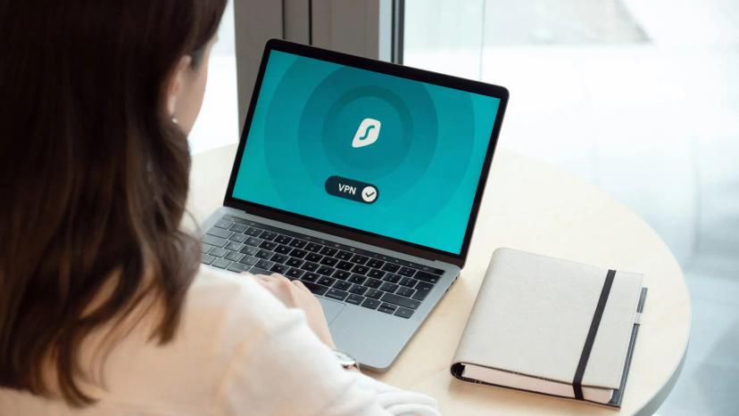 Riscos de segurança na computação em nuvem – como proteger seus dados Mulher de costas utiliza laptop com tela de VPN ativa. Sobre a mesa redonda clara, há um caderno fechado.