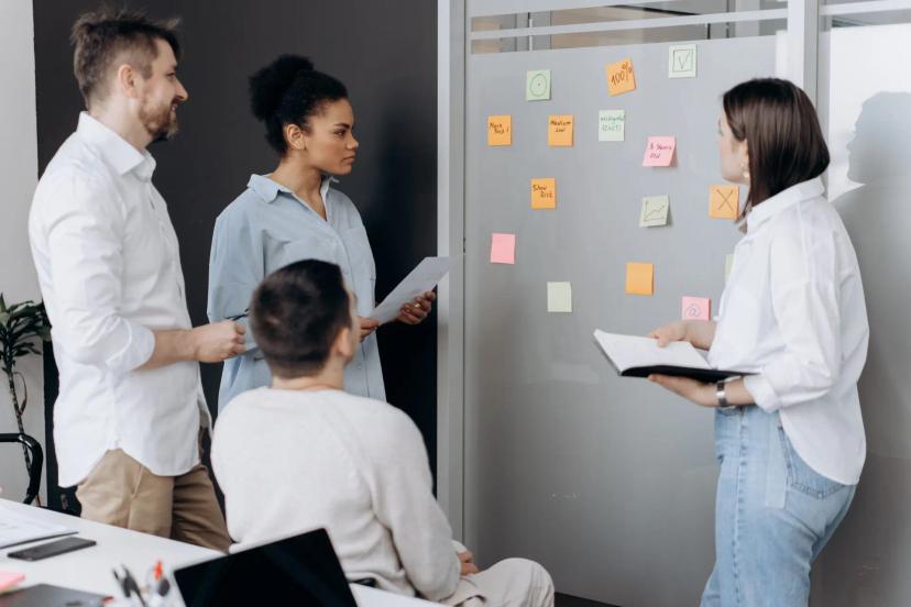 Upskilling e reskilling – o desenvolvimento de competências nas equipes Grupo de quatro profissionais colabora em frente a um quadro de vidro com post-its coloridos em reunião de planeamento.