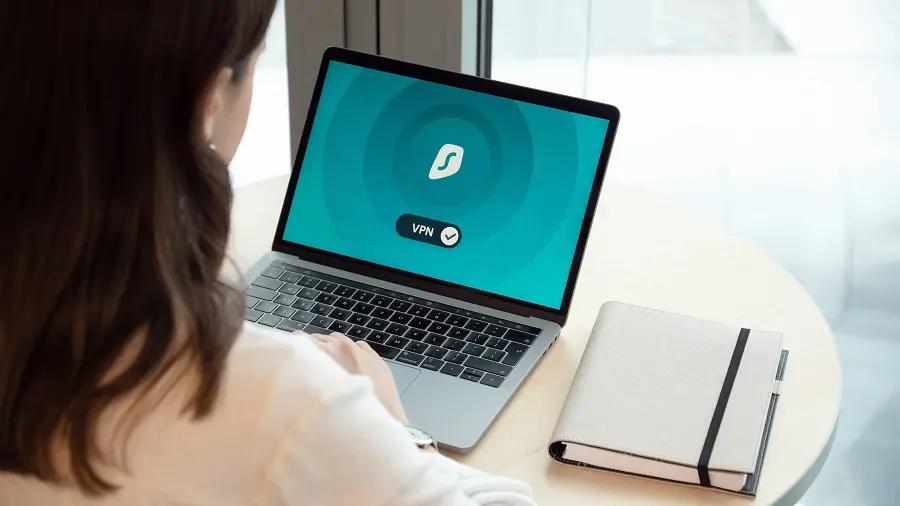Criar software privado por design – prioridade na proteção de dados Mulher a utilizar portátil com ícone de VPN ativa no ecrã, sentada à mesa com um caderno ao lado.
