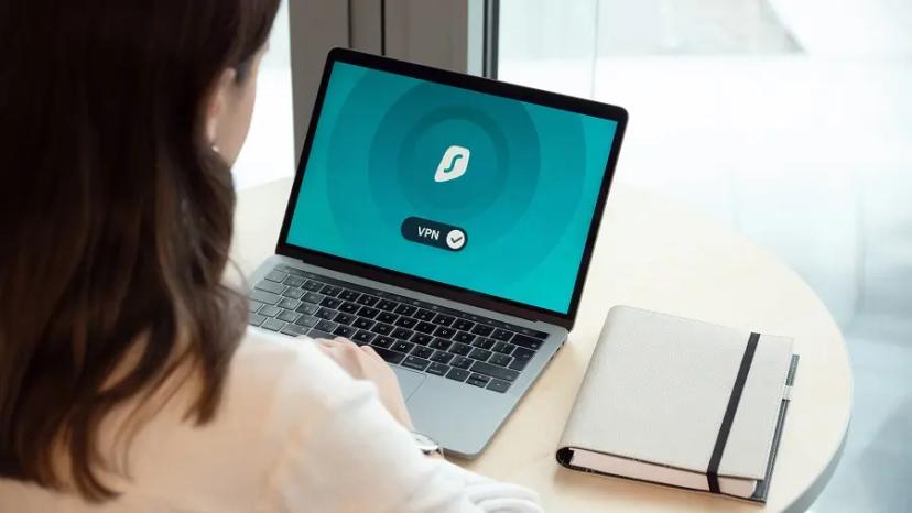Criar software privado por design – prioridade na proteção de dados Mulher a utilizar portátil com ícone de VPN ativa no ecrã, sentada à mesa com um caderno ao lado.