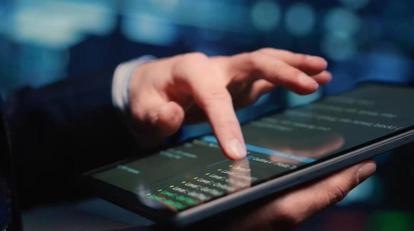 Lei da Informática – como a sua empresa pode beneficiar deste incentivo Mão de uma pessoa a tocar no ecrã de um tablet que exibe linhas de código de programação num ambiente tecnológico.