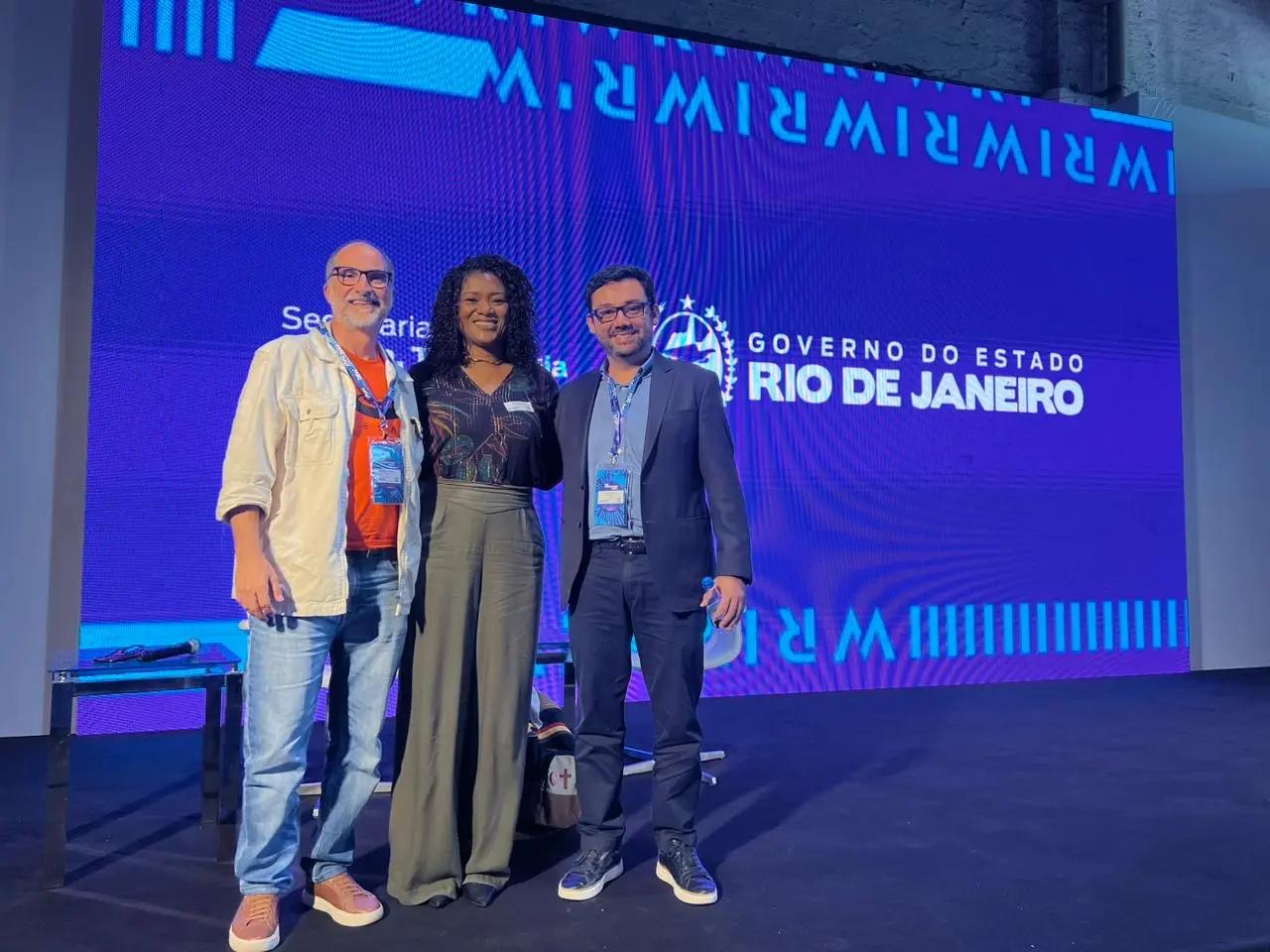 Rio Innovation Week 2023 – reflexões sobre inovação e tecnologia no Brasil Três pessoas sorriem em um palco diante de um telão azul com a marca do Governo do Estado do Rio de Janeiro e Rio Innovation Week.