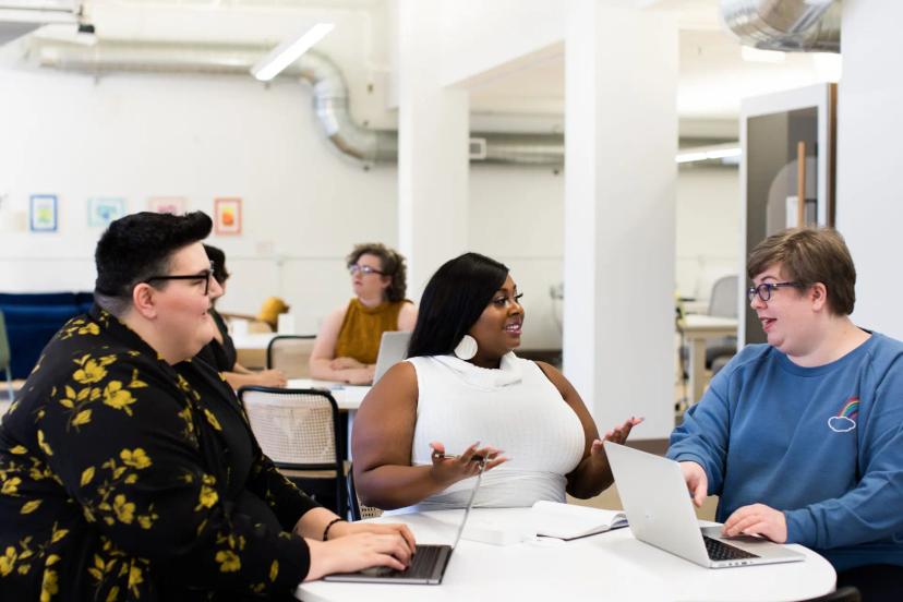 Soft skills na era digital – a importância das habilidades humanas Três mulheres conversam sorridentes ao redor de uma mesa branca com laptops em um escritório moderno e iluminado.