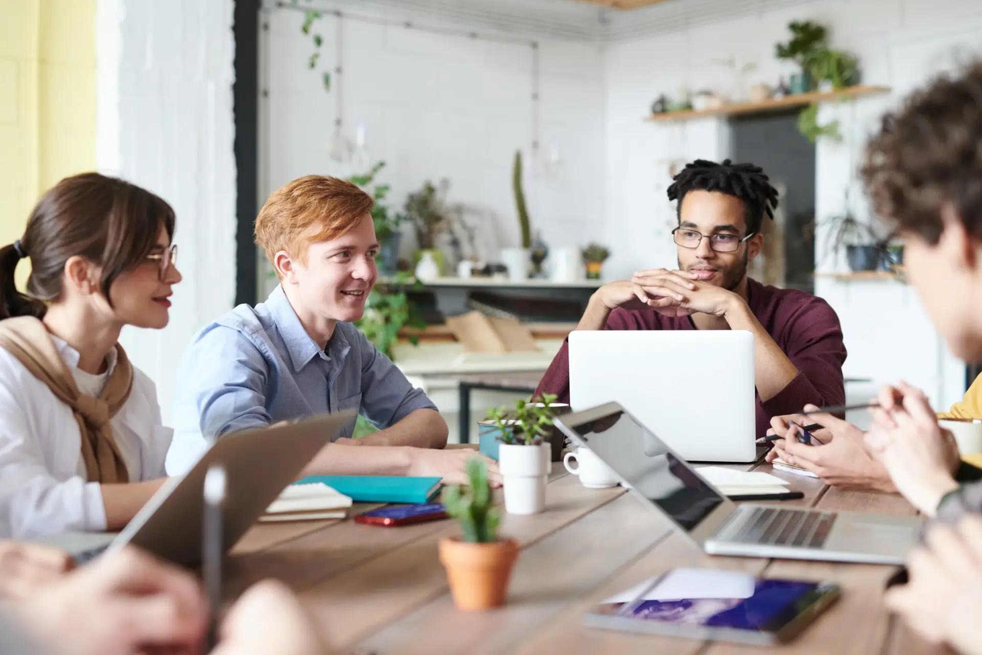 Open Innovation – como acelerar projetos com inovação aberta Grupo de cinco profissionais jovens sorrindo em reunião colaborativa ao redor de mesa com laptops e cadernos em escritório.