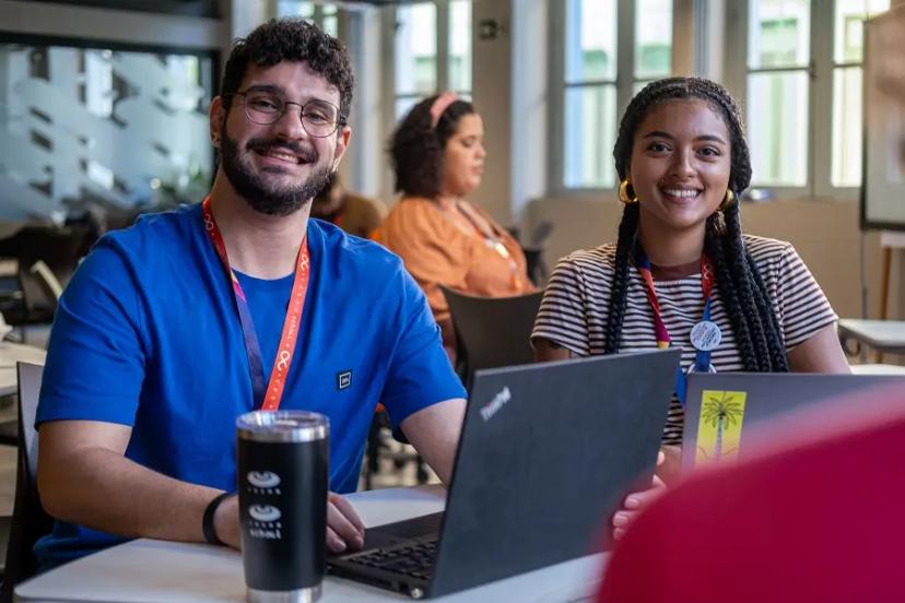 PBL e a colaboração entre alunos em desafios reais de mercado Dois estudantes sorridentes, um homem de azul e uma mulher com camisa de listras, usando laptops em mesa de estudo coletiva.