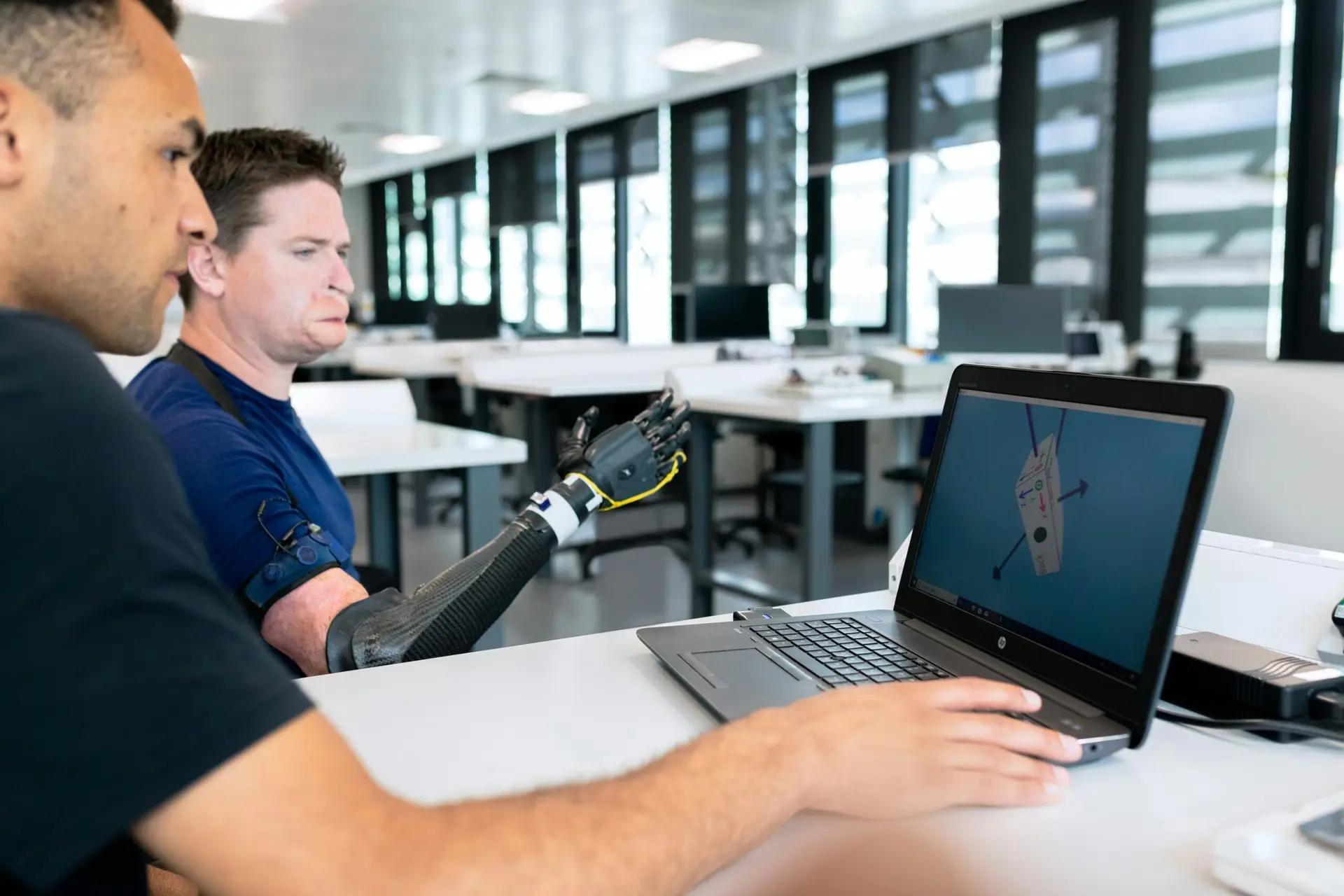 Fontes de fomento à inovação para o desenvolvimento de próteses biônicas Dois homens em laboratório analisando modelo 3D em notebook enquanto um deles testa uma prótese de braço robótico preto.
