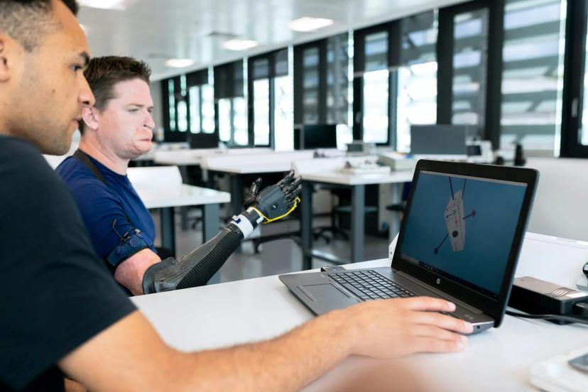 Fontes de fomento à inovação para o desenvolvimento de próteses biônicas Dois homens em laboratório analisando modelo 3D em notebook enquanto um deles testa uma prótese de braço robótico preto.