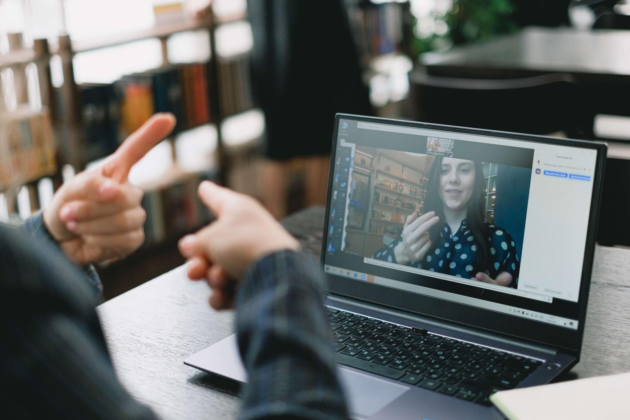 pexels-shvets-production-7516363 Close-up of hands gesturing during a video call on a laptop. A woman in a polka dot shirt is visible on the screen.  The setting appears to be a cafe or library.