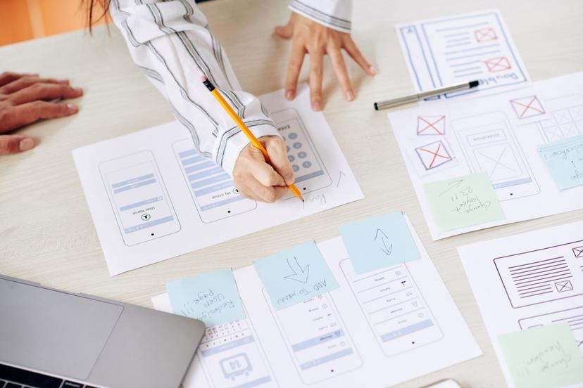 58acbbe9-67a6-9981-eefd-1df72938 Duas designers revisam esboços de protótipos de aplicativos para celular, anotando melhorias em papeis com caneta e post-its.