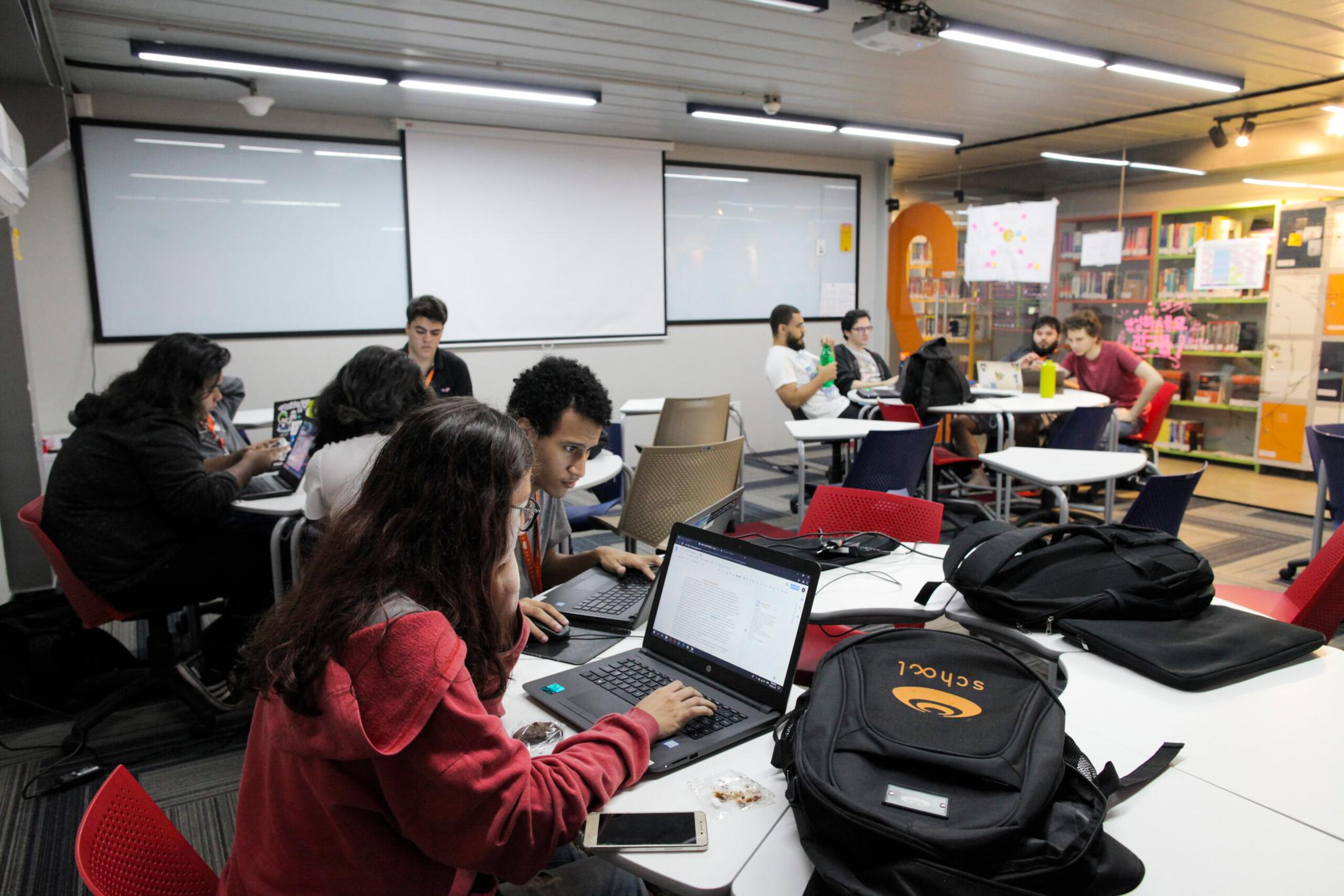 A-revolucao-de-aprender-constantemente-–-CESAR Um grupo de estudantes trabalha em laptops em uma sala de aula moderna e bem iluminada, com mesas, cadeiras e prateleiras de livros ao fundo.