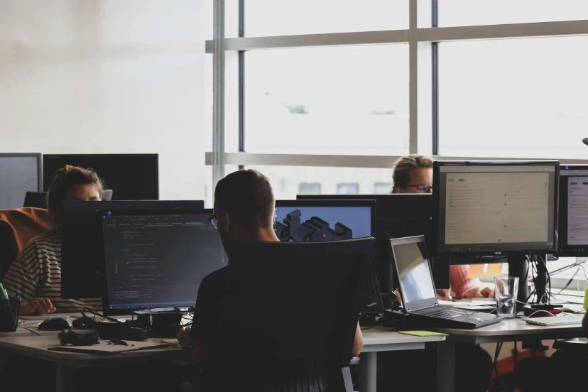 IA Generativa e miniaturização de modelos em ambiente de desenvolvimento Três desenvolvedores trabalhando em computadores com linhas de código e gráficos em escritório iluminado com grandes janelas.