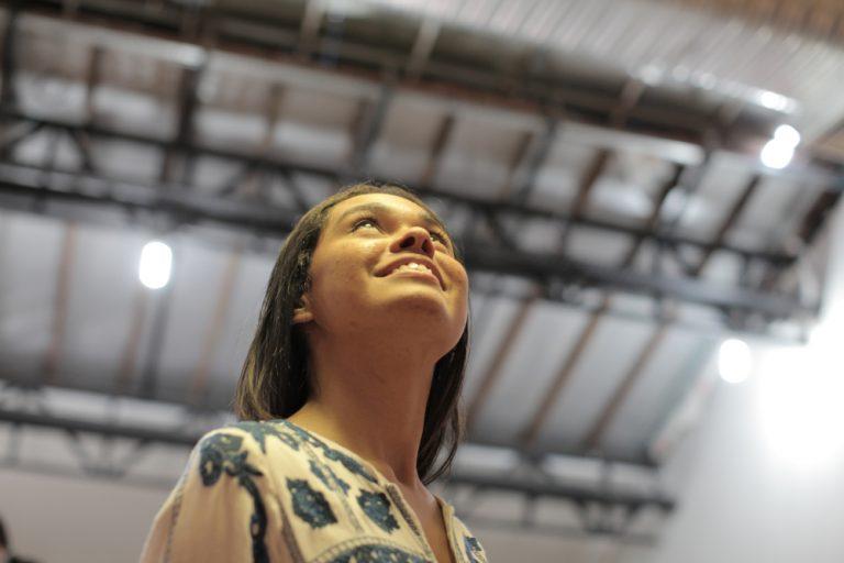 LGPD-garante-ao-titular-de-dados-pessoais-o-direito-de-administra-los_-conheca-as-novas-diretrizes-–-CESAR Uma jovem de cabelos castanhos escuros com uma blusa estampada, olhando para cima com um sorriso e expressão feliz, contra o fundo desfocado de um teto de madeira e vigas de metal.