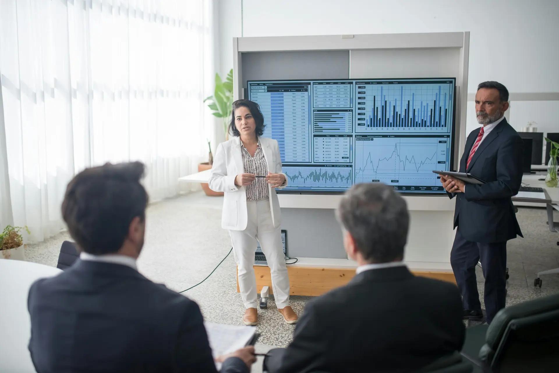 Papel do TRL em inovações de sucesso Executiva de branco apresenta dashboard com gráficos em tela digital para três investidores em sala de reunião corporativa.