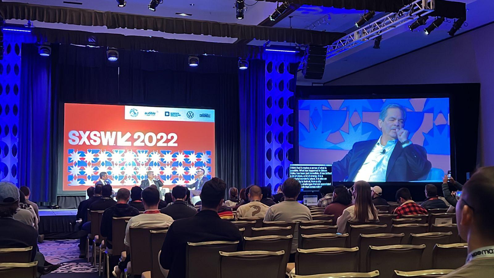 WhatsApp-Image-2022-03-13-at-13.35.34 Painel SXSW 2022 com palestrantes no palco e uma grande tela exibindo um homem falando para uma plateia sentada em um auditório.