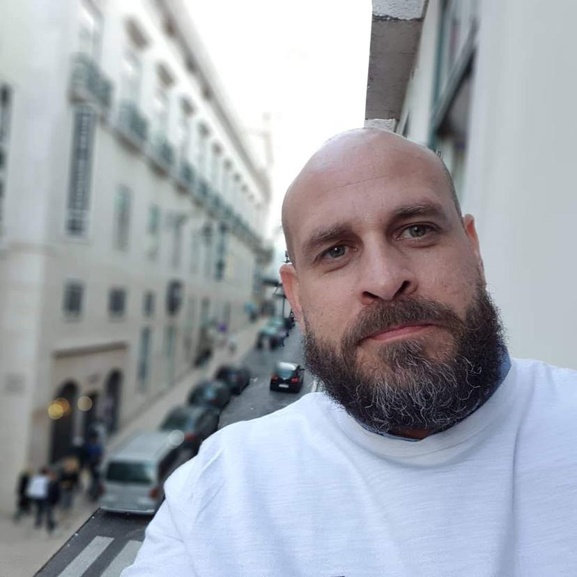 WhatsApp-Image-2022-06-23-at-19.02.31 Homem careca com barba grisalha usando uma blusa branca, em um close-up, com uma rua da cidade desfocada ao fundo.