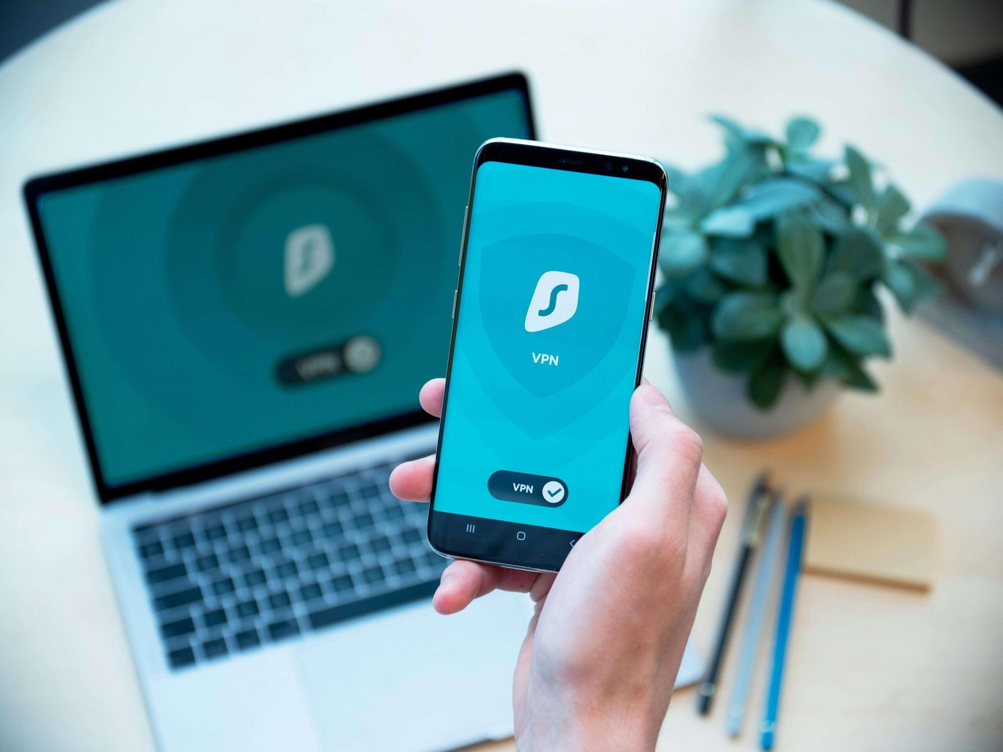 dan-nelson-hIz3vbUbOoI-unsplash (1) Close-up of a hand holding a smartphone displaying a VPN app interface, with a laptop showing a similar VPN interface in the background.