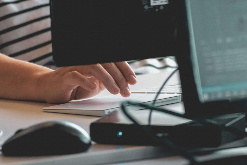 Uma mão usando um trackpad de laptop sobre uma mesa, com um mouse de computador preto e um monitor desfocado em primeiro plano.