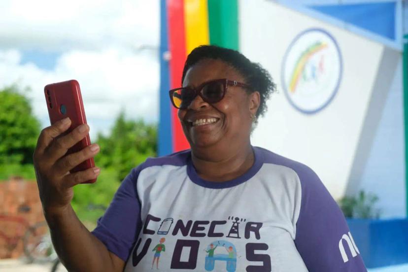 Inclusão Digital em Lagoa de Itaenga – Formação de Jovens e Idosos Mulher negra sorridente usa óculos escuros e segura celular vermelho. Ela veste camisa branca com a frase Conectar Vidas.