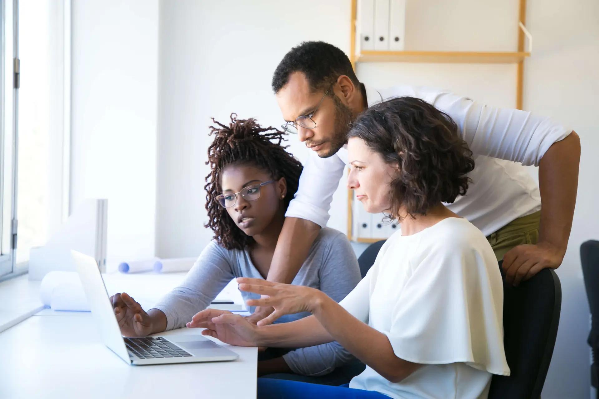 Integração de TI na educação para preparar profissionais do futuro Três profissionais, duas mulheres e um homem, concentrados analisando dados em um laptop branco em escritório iluminado.