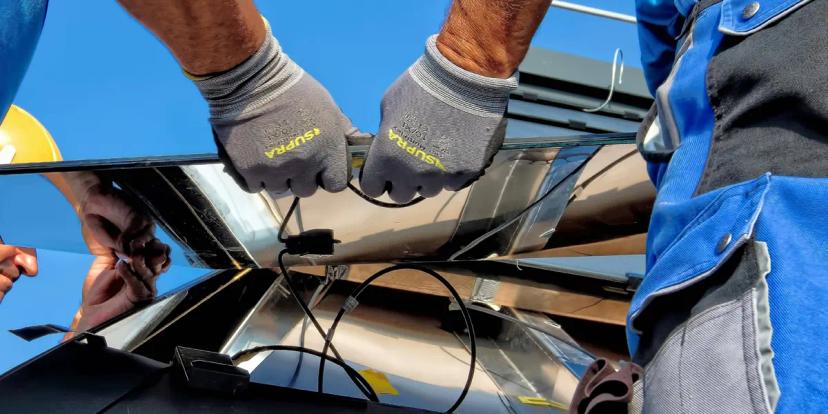 Projetos de P&D no setor de energia Duas pessoas de luvas cinzas conectando cabos sob painel solar. Um técnico de capacete amarelo aparece no reflexo da placa.