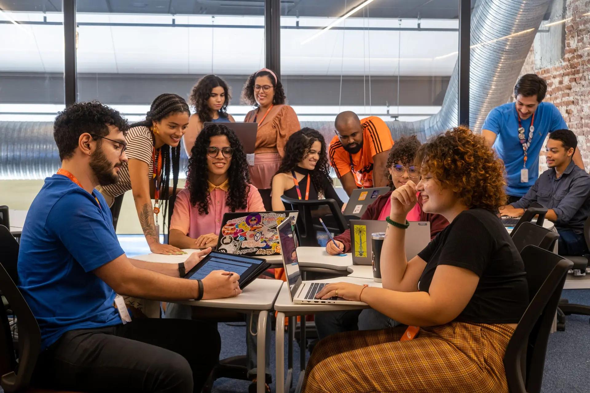 Competências digitais e inclusão em ambiente de aprendizado colaborativo Grupo diverso de jovens em sala moderna utiliza laptops e tablets, sorrindo e interagindo ao redor de mesas brancas.